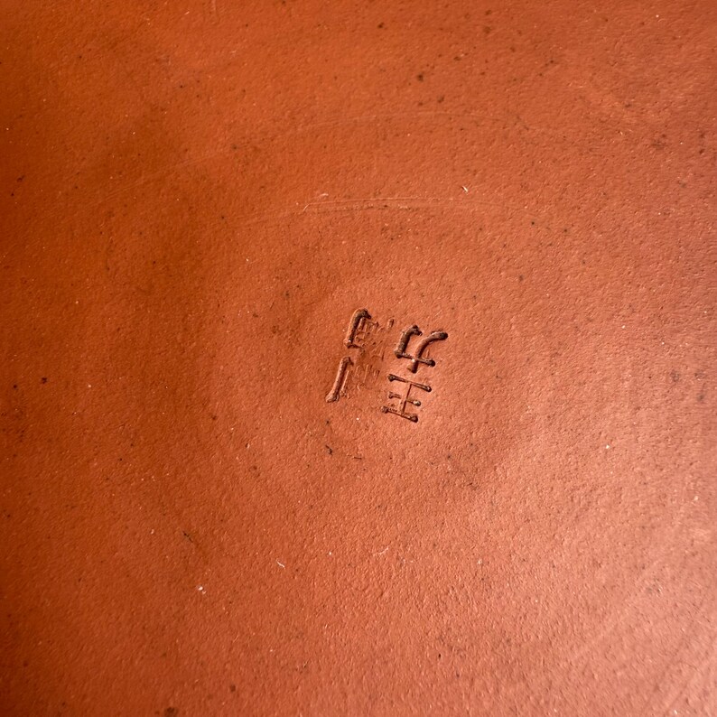 May include: Close-up of a terracotta-colored ceramic surface with stamped Japanese characters. The surface is a warm, earthy tone, and the characters are slightly darker, creating a subtle contrast. The texture appears smooth.