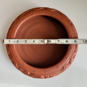 May include: A terracotta-colored, round ceramic dish with a decorative carved border. The dish is approximately 9 inches in diameter, as measured by a ruler. The rim features a repeating pattern of circles and carved designs.
