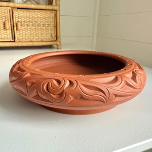 May include: A terracotta-colored, round planter with an intricate carved floral design around the rim. The planter has a wide, shallow bowl shape and a raised base. The carved details add texture and visual interest to the piece.