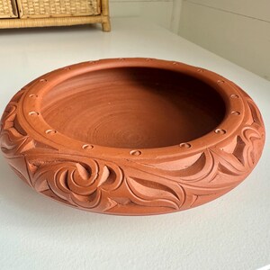 May include: A terracotta-colored, round bowl with carved floral and geometric patterns around the rim. The bowl has a wide, shallow interior and a series of small, evenly spaced holes near the top edge.
