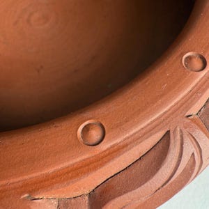 May include: Close-up of a terracotta-colored ceramic pot with a circular indentation and decorative carved details. The pot's rim features two small, evenly spaced circular indentations. The design includes a carved, leaf-like pattern.