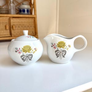 May include: A white ceramic sugar bowl with a lid and a matching creamer. Both pieces are decorated with a yellow flower, gray bow, and floral accents. The set is designed for serving sugar and cream.