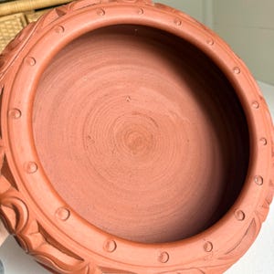 May include: A terracotta-colored, round ceramic bowl with a wide rim. The rim features a repeating pattern of carved floral designs and small circular indentations. The interior of the bowl has a textured, concentric circle pattern.
