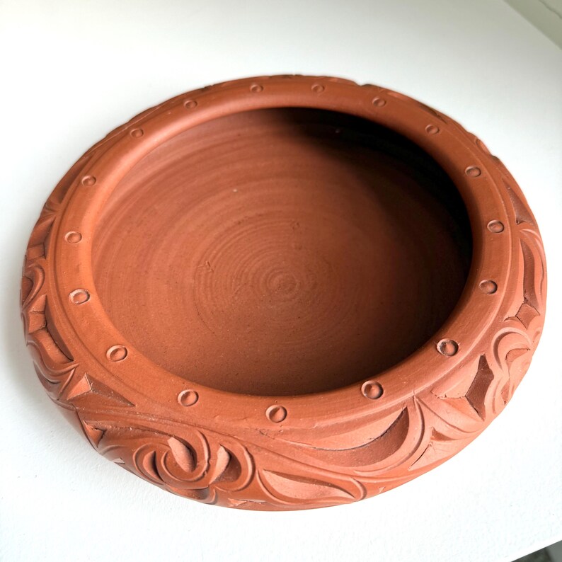May include: A round, terracotta-colored ceramic bowl. The bowl has a wide, shallow design with a textured interior. The rim features a series of small, evenly spaced circles and carved floral patterns. The bowl is likely handmade and has a rustic aesthetic.