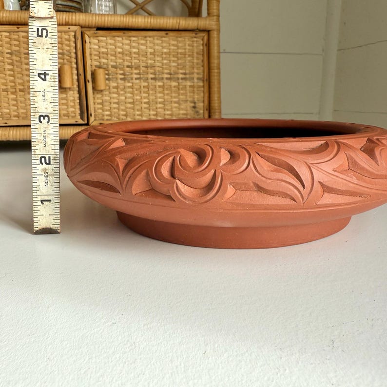 May include: A terracotta-colored, low, round planter with carved floral designs around the rim. The planter is approximately 2 inches (5 cm) tall and has a wide, shallow bowl shape. The base is a slightly darker shade of the same terracotta color.
