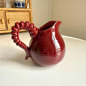 May include: A maroon ceramic pitcher with a unique, textured handle. The handle is a series of connected, rounded shapes. The pitcher has a wide spout and a ribbed body. The pitcher is on a white surface.