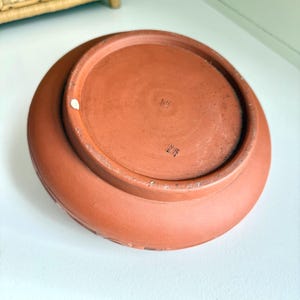 May include: A terracotta-colored ceramic plate, viewed from the underside, showcasing a circular design. The plate has a wide, slightly raised rim with a subtle pattern. The base features stamped markings and a smooth, unglazed surface.