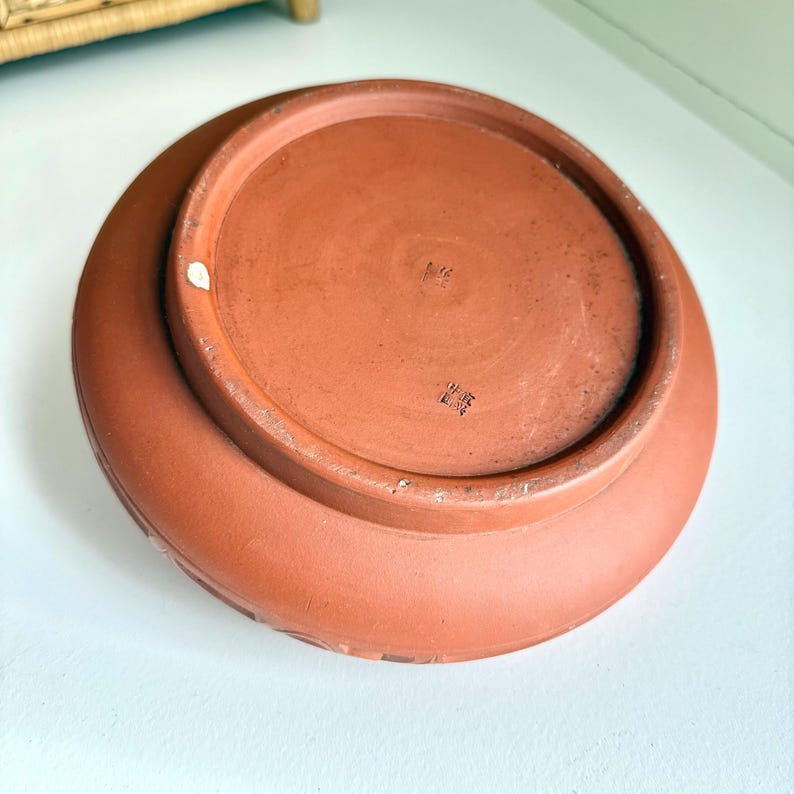 May include: A terracotta-colored ceramic plate, viewed from the underside, showcasing a circular design. The plate has a wide, slightly raised rim with a subtle pattern. The base features stamped markings and a smooth, unglazed surface.