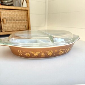 May include: A vintage brown Pyrex divided casserole dish with a clear glass lid. The dish features gold-colored decorative patterns of birds, wheat, and other motifs. The dish is oval-shaped and has a clear glass lid.
