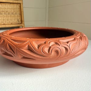 May include: A terracotta-colored, low, round planter with a wide rim. The rim features a carved floral design. The planter sits on a white surface. The interior of the planter is a darker shade of terracotta.