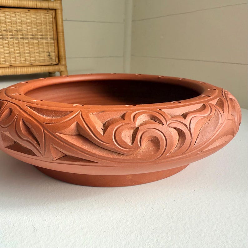 May include: A terracotta-colored, low, round planter with a wide rim. The rim features a carved floral design. The planter sits on a white surface. The interior of the planter is a darker shade of terracotta.