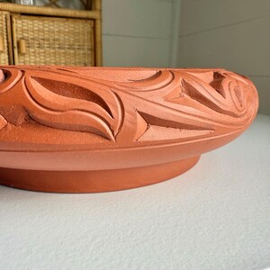 May include: A terracotta-colored, round planter with a detailed, carved floral design around the rim. The planter has a wide base and a slightly curved lip. The carved pattern features stylized leaves and geometric shapes.