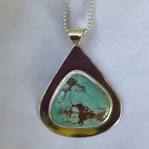 May include: A teardrop-shaped silver pendant with a turquoise stone set in the center. The stone has a mottled pattern of brown and green.
