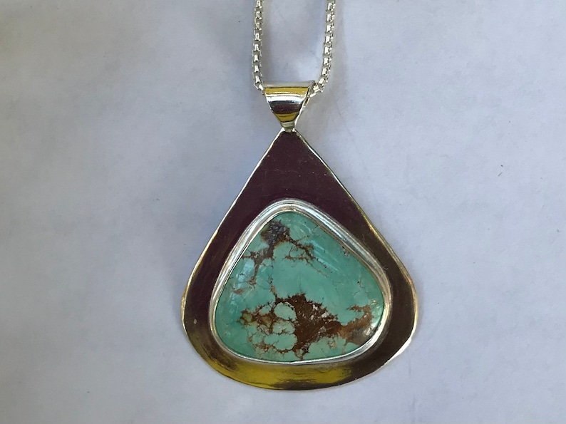 May include: A teardrop-shaped silver pendant with a turquoise stone set in the center. The stone has a mottled pattern of brown and green.
