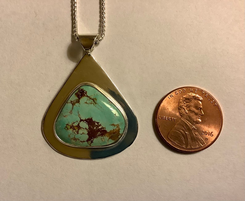 May include: A silver pendant with a turquoise stone set in a teardrop shape. The pendant is about the size of a US penny.