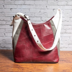 May include: A patchwork-style shoulder bag in burgundy, white, and gray. The bag features a white strap with silver-tone hardware and a rectangular shape. The bag is made of a leather-like material and has a unique design.