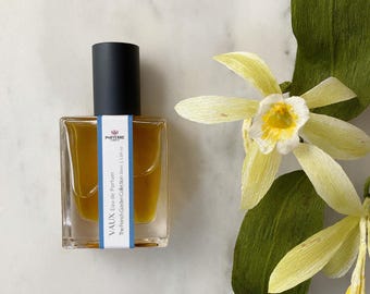 May include: A square glass perfume bottle with a black cap, filled with a golden liquid. The label reads "VAUX Eau de Parfum" and "The French Garden Collection 30ml | 1.0fl oz." Beside it are paper flowers with yellow petals and green leaves.