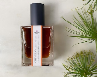 May include: A square perfume bottle with a dark navy cap and a label that reads "CHEVERNY Eau de Parfum". The bottle contains a brown liquid and is part of "The French Garden Collection 30ml 1.0 fl oz". Green foliage is in the background.