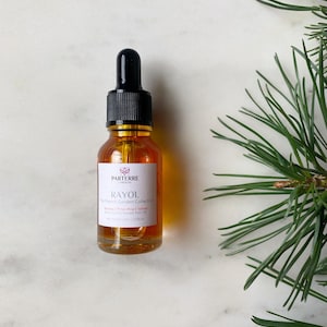 May include: A small amber glass bottle with a black dropper cap. The label reads "PARTERRE GARDENS RAYOL The French Garden Collection". The bottle contains a clear, golden liquid. A sprig of green pine needles is in the background.