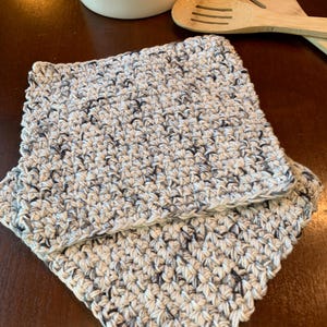 May include: Two crocheted dishcloths in a white and gray speckled yarn. The dishcloths are folded on top of each other.