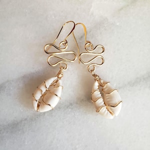 May include: A pair of gold-toned wire-wrapped dangle earrings featuring white cowrie shells. Each earring has a decorative gold wire loop design at the top, connected to a single cowrie shell that is securely encased in thin gold wire wrapping.
