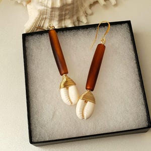 May include: A pair of handcrafted earrings featuring a natural seashell design. The earrings have a brown wooden rod, a white seashell, and gold-colored wire wrapping. The earrings are displayed in a black box with white padding, with a seashell in the background.