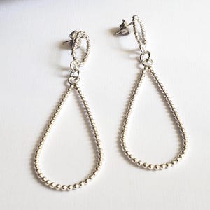 Pode incluir: Um par de brincos prateados em forma de gota. Cada brinco apresenta um design de contas, com um pequeno laço na parte superior e uma forma de gota maior feita de pequenas contas. Os brincos são exibidos em uma superfície branca.