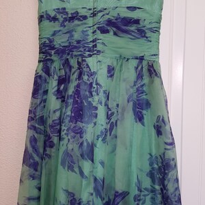 May include: A sleeveless, knee-length dress in shades of green and blue, hanging on a black hanger. The dress features a V-neck and a gathered waist. The fabric is a sheer material with a floral pattern in blue and green.