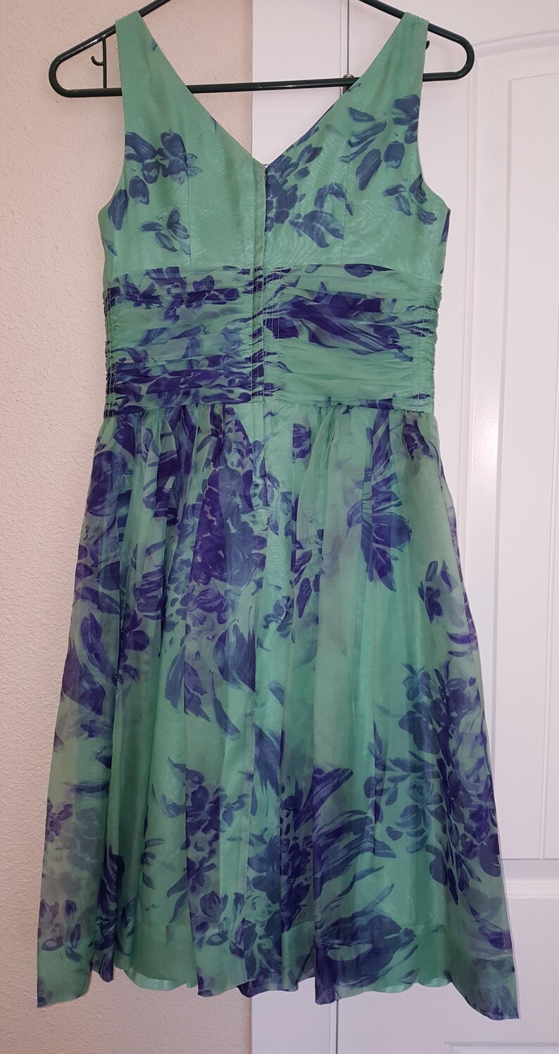 May include: A sleeveless, knee-length dress in shades of green and blue, hanging on a black hanger. The dress features a V-neck and a gathered waist. The fabric is a sheer material with a floral pattern in blue and green.