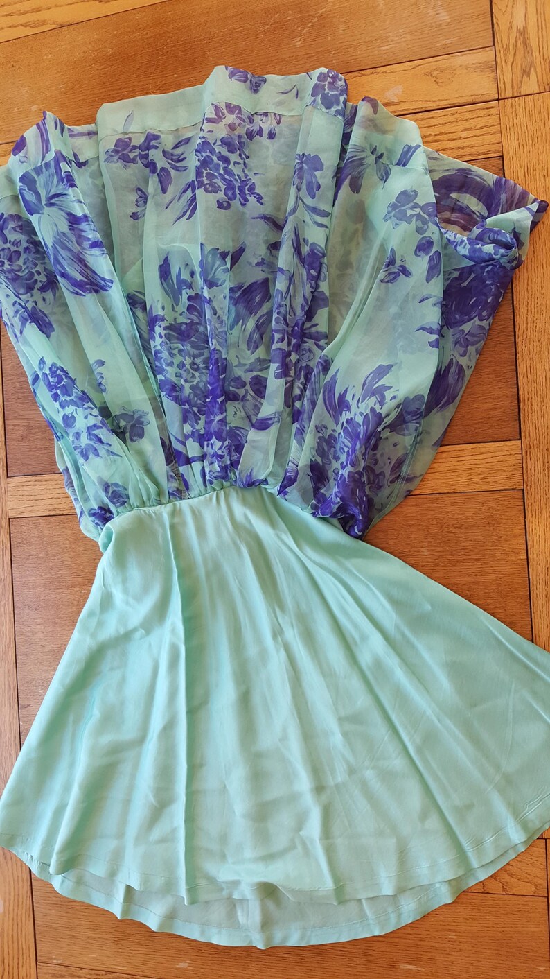 May include: A mint green dress with a sheer overlay featuring a purple floral pattern. The dress has a gathered waist and a flowing skirt, creating a soft, feminine silhouette. The fabric appears lightweight and suitable for warm weather.