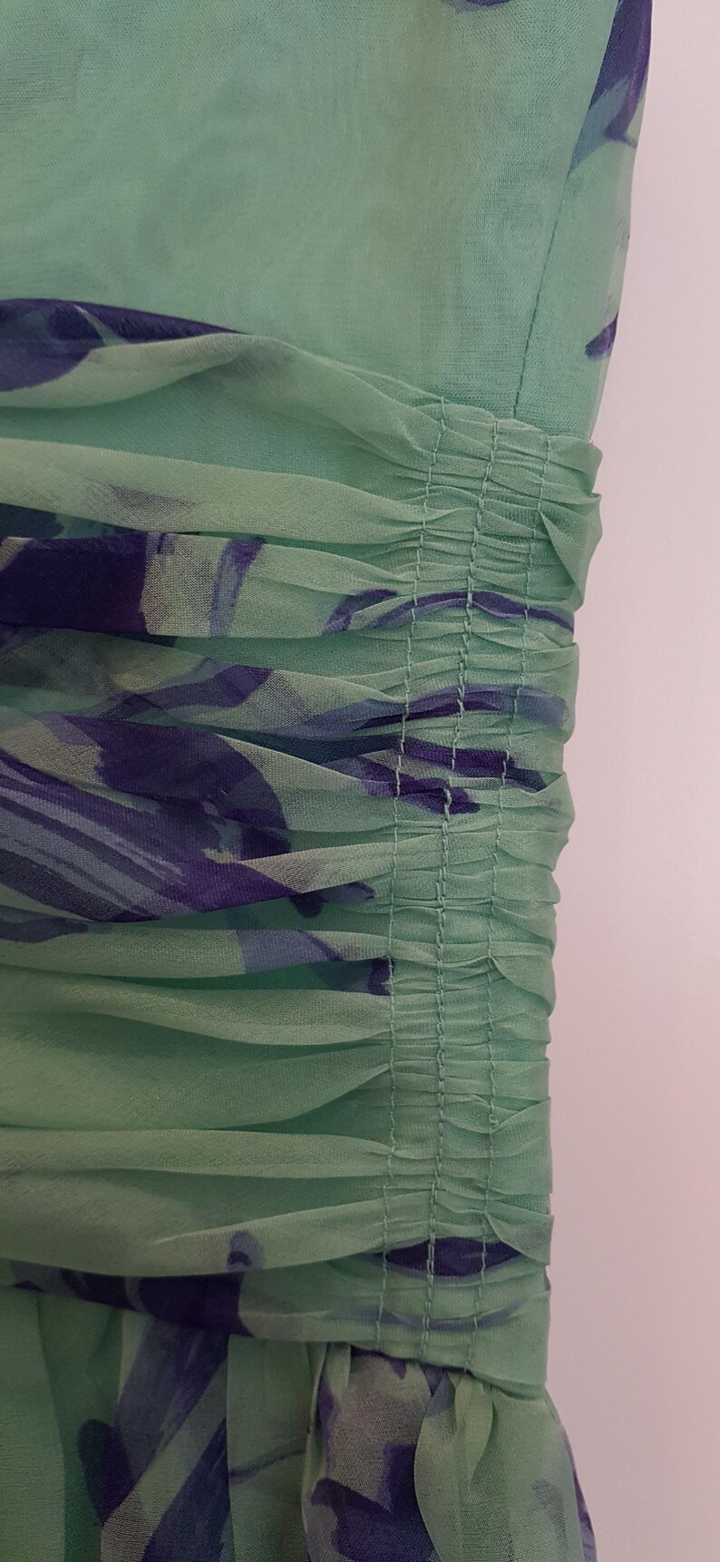May include: Close-up of a mint green dress with a floral pattern in shades of purple and blue. The fabric is sheer and gathered, creating a textured effect. The dress appears to be made of a lightweight material.