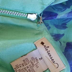 May include: Close-up of a teal-colored garment with a zipper and a tag. The tag reads "elevenses" and lists the materials as 100% silk shell and 100% viscose lining. The garment is size 2 and made in India.