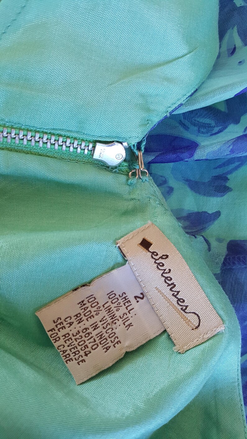 May include: Close-up of a teal-colored garment with a zipper and a tag. The tag reads "elevenses" and lists the materials as 100% silk shell and 100% viscose lining. The garment is size 2 and made in India.