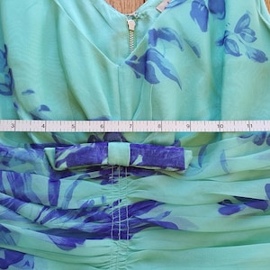 May include: A sleeveless, light green dress with a V-neck and a bow detail. The dress features a blue floral pattern and a gold zipper. A measuring tape shows the dress is approximately 13 inches wide.