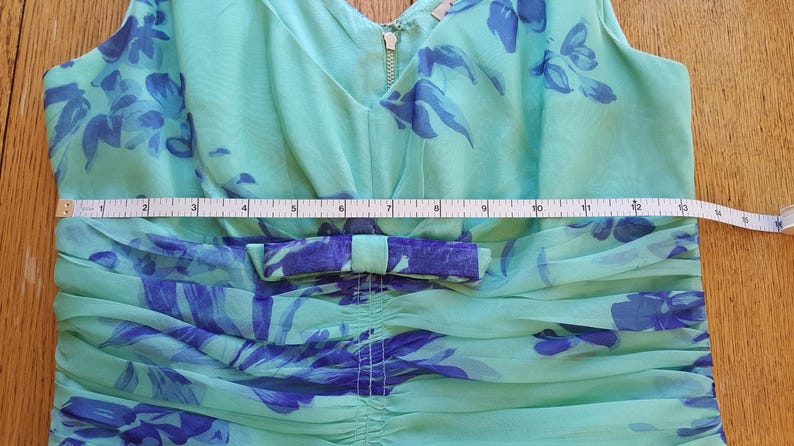May include: A sleeveless, light green dress with a V-neck and a bow detail. The dress features a blue floral pattern and a gold zipper. A measuring tape shows the dress is approximately 13 inches wide.