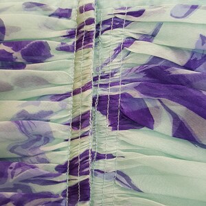 May include: Close-up of a mint green fabric with a floral pattern in shades of purple. The fabric is gathered and ruched, with vertical seams running down the center. The overall effect is soft and flowing, suggesting a dress or other garment.