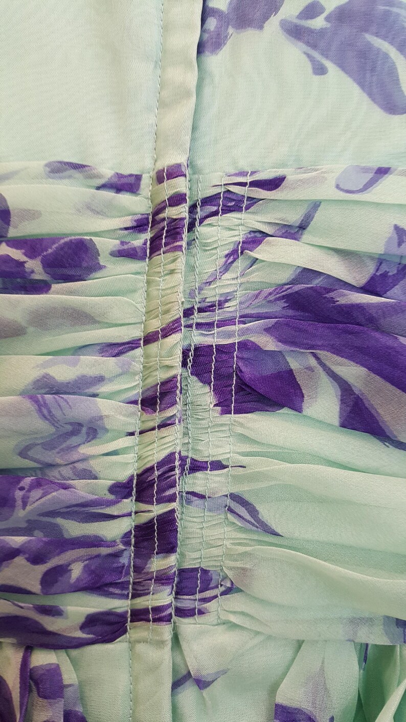 May include: Close-up of a mint green fabric with a floral pattern in shades of purple. The fabric is gathered and ruched, with vertical seams running down the center. The overall effect is soft and flowing, suggesting a dress or other garment.