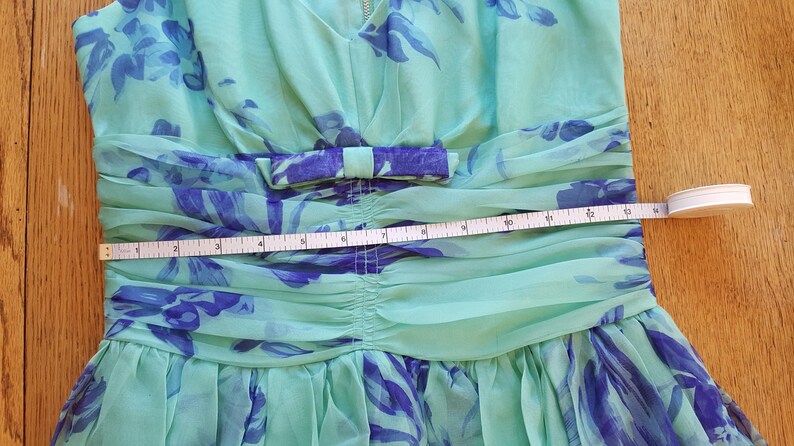 May include: A light green dress with a blue floral pattern and a bow at the waist. A measuring tape is placed across the waist of the dress, showing a measurement of approximately 10 inches.