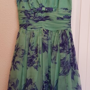 May include: A sleeveless, knee-length dress in shades of green and blue, hanging on a black hanger. The dress features a V-neck, a ruched waist with a bow, and a floral pattern. The sheer fabric drapes gracefully.