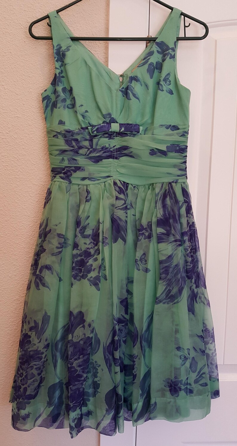 May include: A sleeveless, knee-length dress in shades of green and blue, hanging on a black hanger. The dress features a V-neck, a ruched waist with a bow, and a floral pattern. The sheer fabric drapes gracefully.