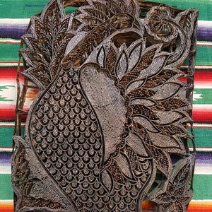 May include: A rectangular dark brown wooden block print stamp featuring an intricate carved design of a large bird with detailed plumage, curved wings, and floral elements. The surface displays fine metal wire inlays forming the patterns.