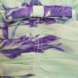 May include: Close-up of a mint green dress with a purple floral pattern. The dress features a bow detail at the neckline and gathered fabric. The fabric is sheer and lightweight, ideal for warm weather.