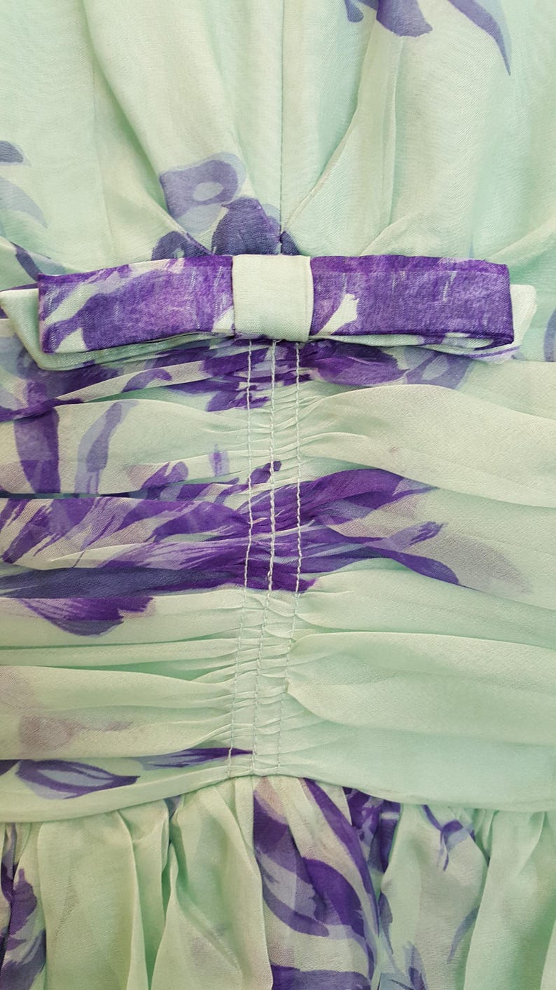 May include: Close-up of a mint green dress with a purple floral pattern. The dress features a bow detail at the neckline and gathered fabric. The fabric is sheer and lightweight, ideal for warm weather.
