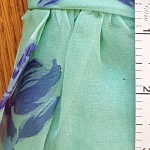 May include: Close-up of a light green fabric with a blue floral pattern. The fabric is gathered at the waist and appears to be approximately 2 inches wide, as measured by a ruler. The fabric is likely used for clothing or accessories.