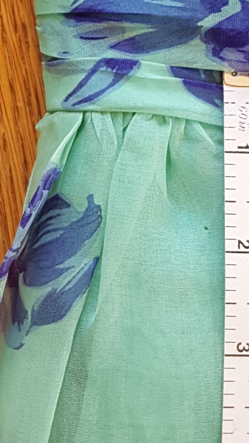 May include: Close-up of a light green fabric with a blue floral pattern. The fabric is gathered at the waist and appears to be approximately 2 inches wide, as measured by a ruler. The fabric is likely used for clothing or accessories.