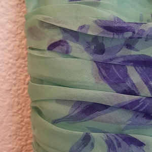 May include: Close-up of a mint green dress with a floral pattern in shades of blue and purple. The fabric is gathered at the waist, creating a textured effect. The dress appears to be made of a lightweight, flowing material.