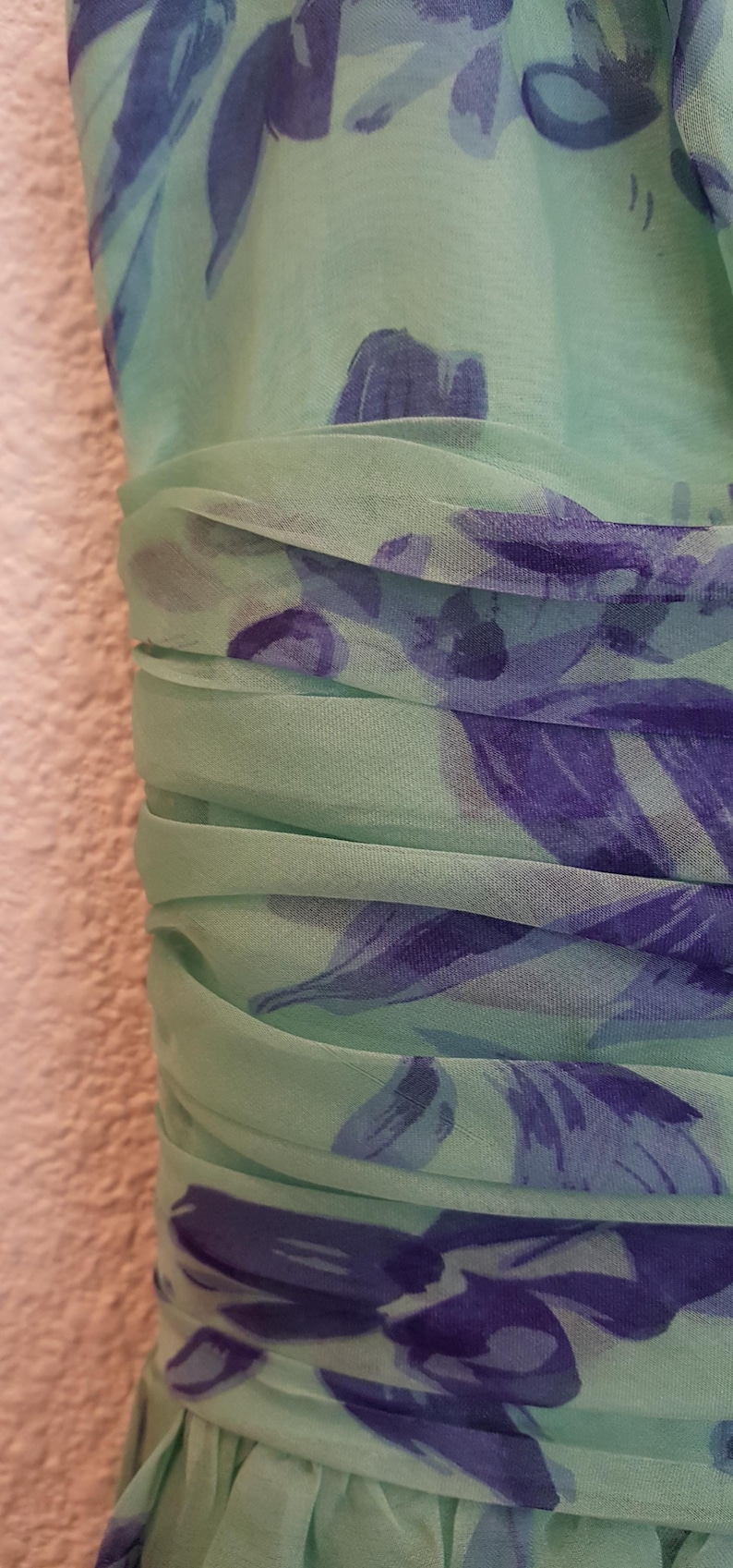 May include: Close-up of a mint green dress with a floral pattern in shades of blue and purple. The fabric is gathered at the waist, creating a textured effect. The dress appears to be made of a lightweight, flowing material.