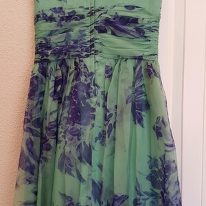 May include: A sleeveless, knee-length dress in a soft green hue, adorned with a floral pattern in shades of blue and purple. The dress features a ruched bodice and a flowing skirt, hanging on a black hanger.