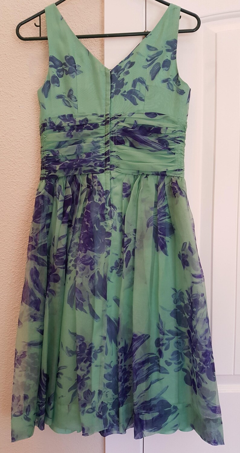 May include: A sleeveless, knee-length dress in a soft green hue, adorned with a floral pattern in shades of blue and purple. The dress features a ruched bodice and a flowing skirt, hanging on a black hanger.