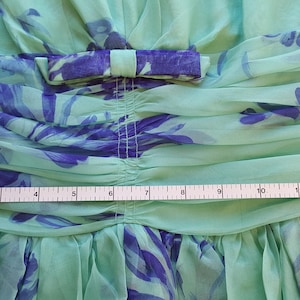 May include: Close-up of a light green dress with blue floral patterns. The dress features a bow at the waist and a gathered bodice. A measuring tape shows a width of approximately 13 inches.